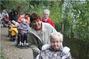 Ehrenamtliche Mitarbeiterinnen und Mitarbeiter machen einen Ausflug mit Bewohnern des Caritas-Altenzentrums St. Josefspflege, die im Rollstuhl sitzen. Sie fahren mit ihnen durch eine Kleingartenanlage
