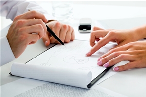 Hände einer Mitarbeiterin zeigen einem Zuhörer, von dem ebenfalls nur die Hände zu sehen sind, auf eine Auflistung in einem Dokument. 