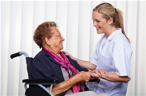 Eine junge Mitarbeiterin der Altenpflege hält die Hände einer älteren Dame im Rollstuhl. Beide zeigen eine fröhliche Miene. 