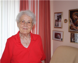 Eine ältere Dame mit Brille und roter Bluse in ihrem Zimmer im Caritas-Altenzentrum St. Josefspflege vor einer Wand mit Fotografien. Wohnen in der St. Josefspflege