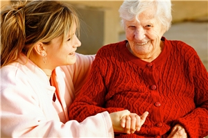 Eine Altenpflegerin sitzt neben einer älteren Dame in einer roten Jacke und hält ihre Hand.