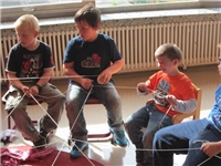 Vier Jungen sitzen auf Stühlen und halten ein Netz aus Fäden in der Hand.