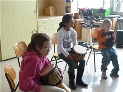 Ein Mädchen und zwei Jungen sitzen mit Trommeln in der Hand auf Stühlen und warten auf ihren Einsatz.