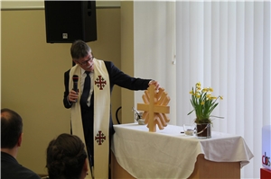 Herr Hundemer stellt das Kreuz vor. Das Kreuz, das im Erdgeschoss des Förderzentrums gesegnet wurde, hat die Form eines Flammenkreuzes, das Logo der Caritas. 