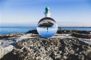 Ein Leuchtturm spiegelt sich in einer runden Glaskugel, im Hintergrund das Meer