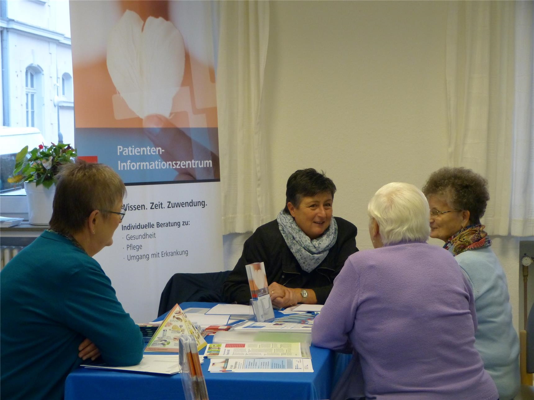 vier Frauen an einem Tisch, im Hintergrund ein Banner mit der Aufschrift „Patienten-Informationszentrum“. (Quartiersmanagement Trier-West/Caritasverband Trier e. V.)