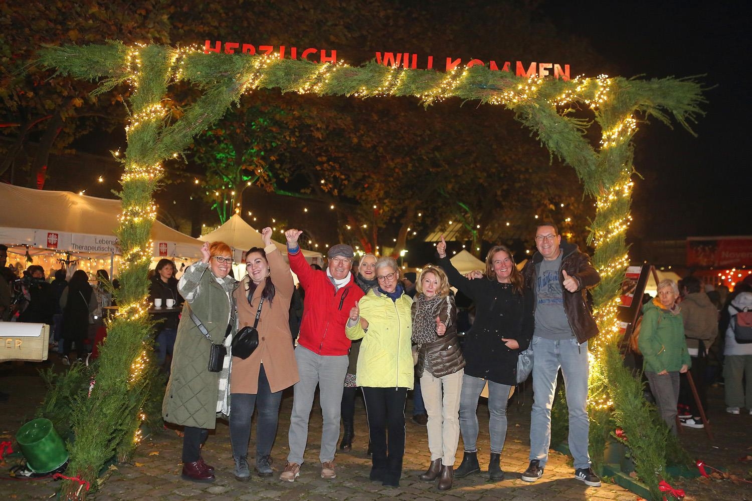 Mit Tannenzweigen und Lichterkette geschmückter Holz-Durchgang vor den Novemberlichtern unter dem eine Gruppe von Menschen steht. 