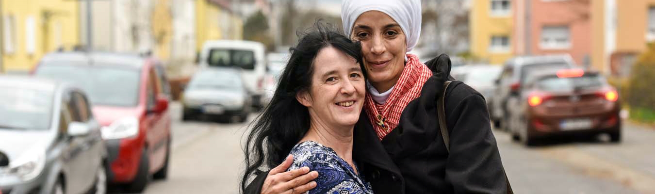 Eine Quartiersarbeiterin und eine Frau mit Kopftuch blicken Arm in Arm in die Kamera