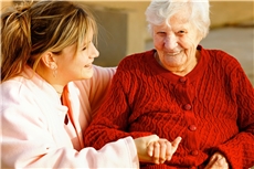 Eine Altenpflegerin sitzt neben einer älteren Dame in einer roten Jacke und hält ihre Hand. / Fotolia