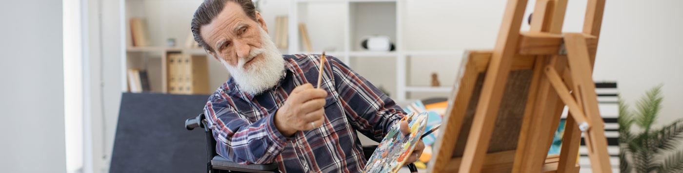 Mann mit weißem Bart im Rollstuhl malt an einer Staffelei