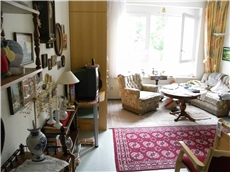 Der Blick in ein Zimmer zeigt links viele persönliche Erinnerungen. Rechts lädt vor dem hellen Fenster ein Sessel mit Sofa und Tisch zum Fernsehen ein.