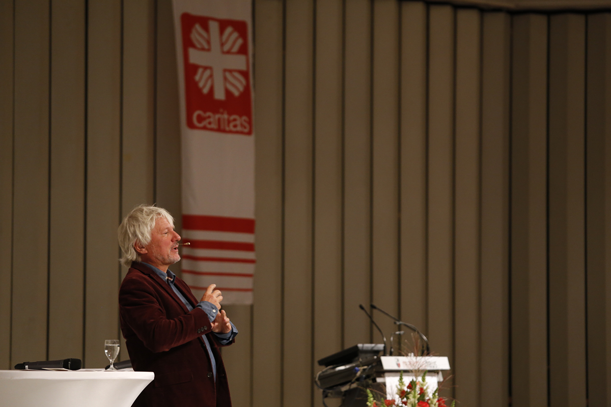  Kabarettist Jürgen Becker steht beim Fachforum'Von der Kunst, ein christliches Unternehmen zu führen' auf einer Bühne. Neben ihm sind ein Pult und eine Caritas-Fahne zu sehen. (Martin Karski)
