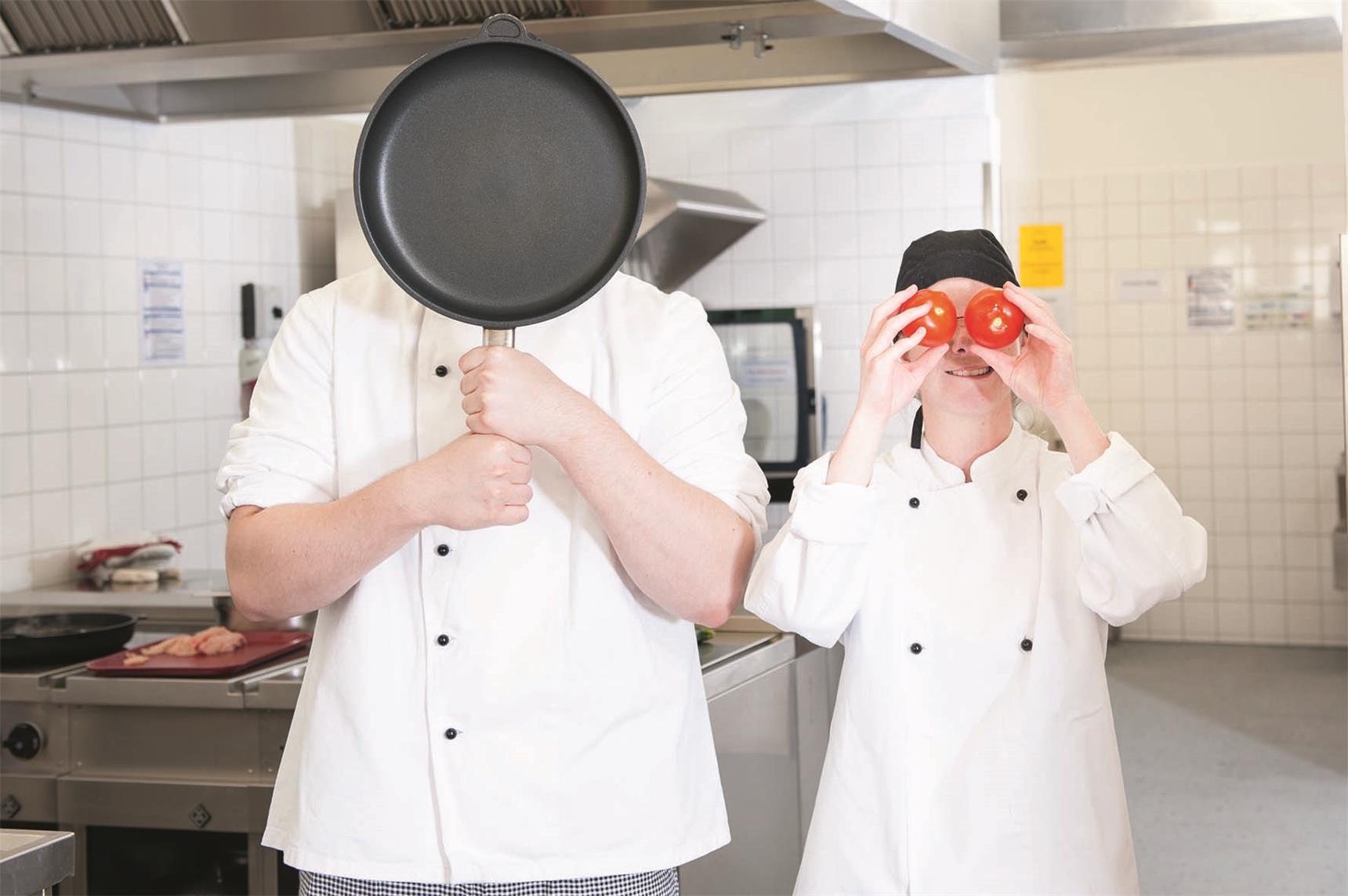 Zwei junge Leute stehen in Schürze in der Küche, einer hält die Pfanne vors Gesicht, die andere zwei Tomaten vor den Augen (Ina Funk)