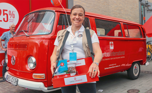Eine Mitarbeiterin steht mit einem Bauchladen vor einem roten VW-Bus von caritas international auf dem Katholikentag 2018 (Caritas)