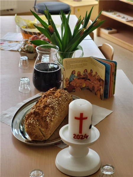Auf einem Tisch stehen ein Brot, eine Karaffe Traubensaft, ein Bilderbuch und eine Osterkerze.
