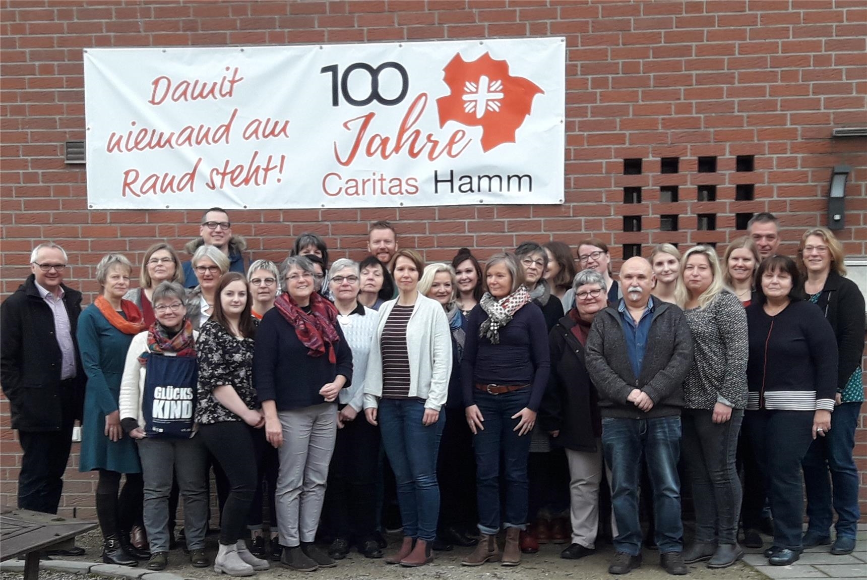 Frauen und Männer stehen als Gruppe zusammen vor einer roten Klinkerwand, an der ein Banner mit "Damit niemand am Rand steht! 100 Jahre Caritas Hamm" steht (Caritas Hamm)