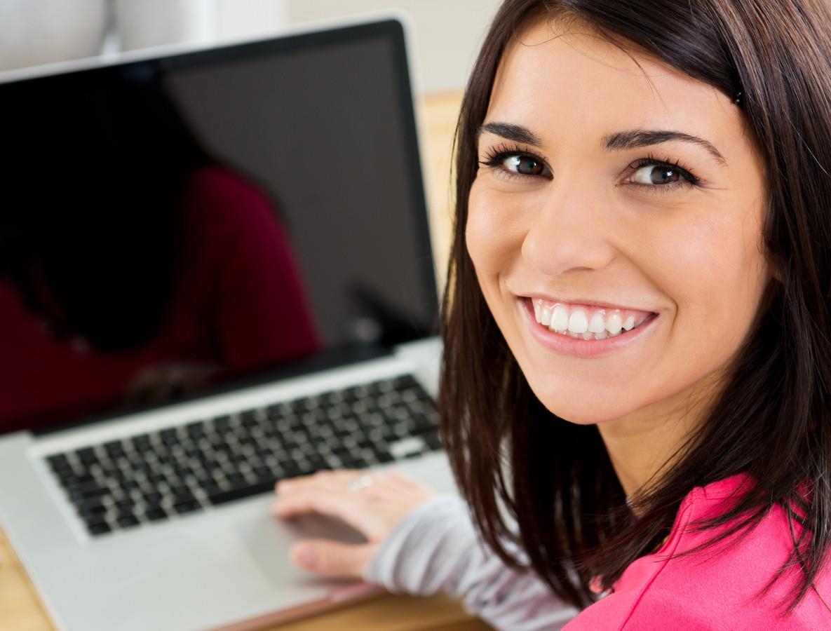Eine junge Frau mit langen dunklen Haaren blickt den Betrachter an. Sie sitzt an einem Laptop, auf dem ihre Hand ruht.