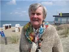 Eine alte Dame steht am Strand, im Hintergrund sind Strandkörbe und die Ostsee zu sehen.