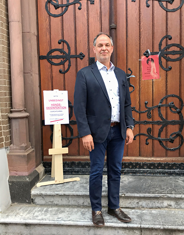 Josef Aretz, der auf der Treppe vor der Tür des Katharina Kasper-Heims in Gangelt steht. Vor der Tür befinden sich zudem zwei Hinweisschilder zur Handdesinfektion. (Foto: Katharina Kasper ViaNobis GmbH)