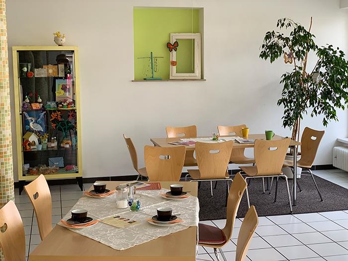 Mehrere Tische mit Stühlen, darauf Kaffeetassen und Zeitungen, ein Benjamini und ein Schrank mit Kunstgewerblichen Artikeln darin (Caritasverband Darmstadt e. V. / Jens Berger)