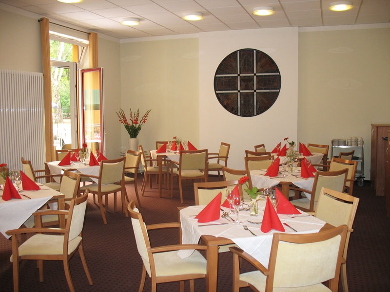 Der mit roten Servietten festlich gedeckte Speisesaal mit rundem Bleiglasornament in der Wand lädt zum Essen ein. 