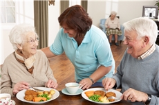 Eine Mitarbeiterin kommt zu einem älteren Ehepaar, das in einer Seniorenwohneinrichtung zu Mittag isst. / Fotolia