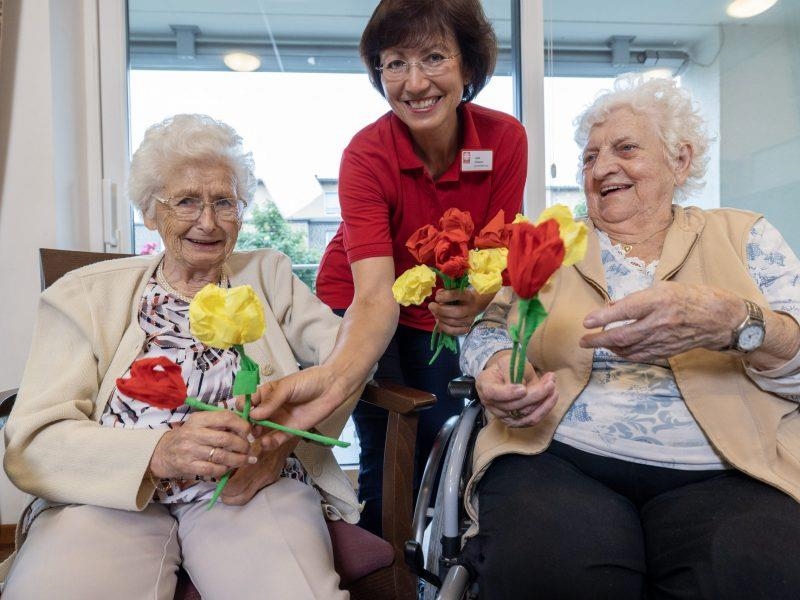 Pflegekraft überreicht zwei Seniorinnen Papierblumen 