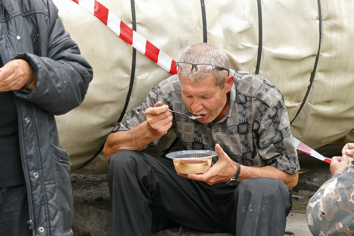 Ein russischer Obdachloser sitzt auf einem Stein und ist eine Suppe aus einer Plastikschale. Um ihn herum sind weitere Obdachlose zu sehen. (Christian Heidrich)