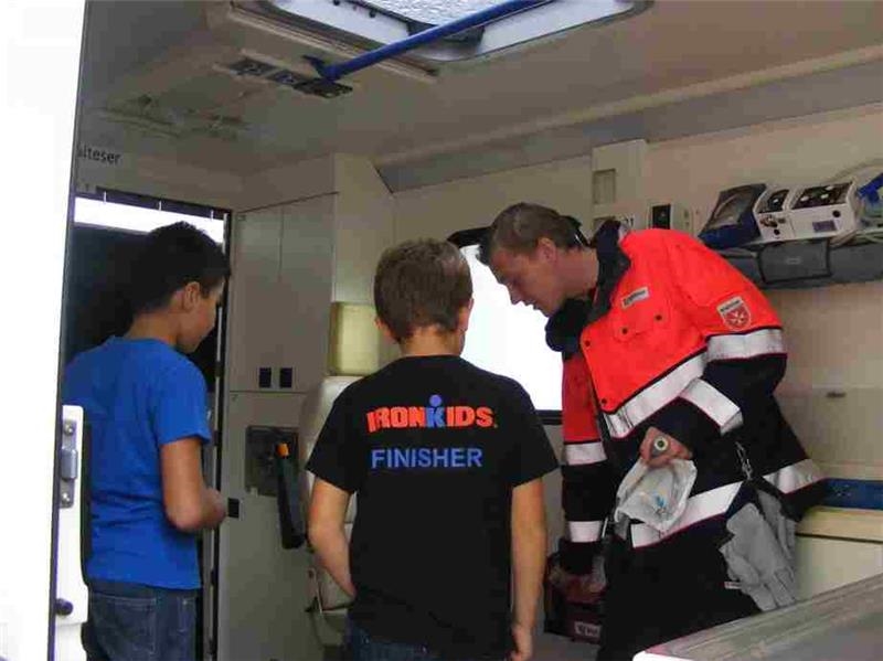 2 Jungen besichtigen einen Rettungswagen. Sie erhalten Erläuterungen von einem Sanitäter in Malteser-Einsatzkleidung.  (Caritasverband Rhein-Hunsrück-Nahe)