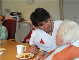 Ein junger Mann mit langen, schwarzen Haaren sitzt bei einer Seniorin und hilft ihr beim Kuchen essen / Foto: Schedlbauer