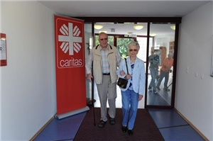 Das Flammenkreuz, das Logo der Caritas, hängt im Eingangsbereich einer Altenpflegeeinrichtung, zwei Besucher, ein Mann und eine Frau, betreten gerade die Einrichtung. 