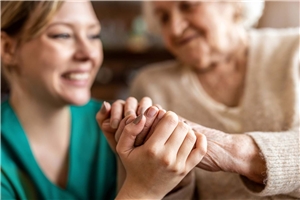 Eine junge Pflegerin hält in Ihren zwei Händen die Hand einer älteren Dame. 