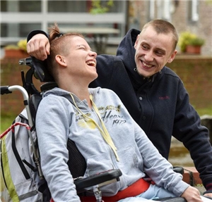 Sozialassistent legt den Arm um Frau mit Behinderung im Rollstuhl, die lacht