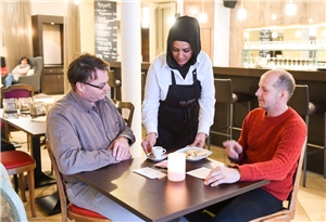 Eine Frau mit Kopftuch bedient zwei Gäste in einem Café