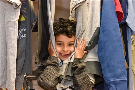Portrait eines kleinen Jungen, der sein Gesicht zwischen den Klamotten eines Kleiderst�nders hervorstreckt und lacht