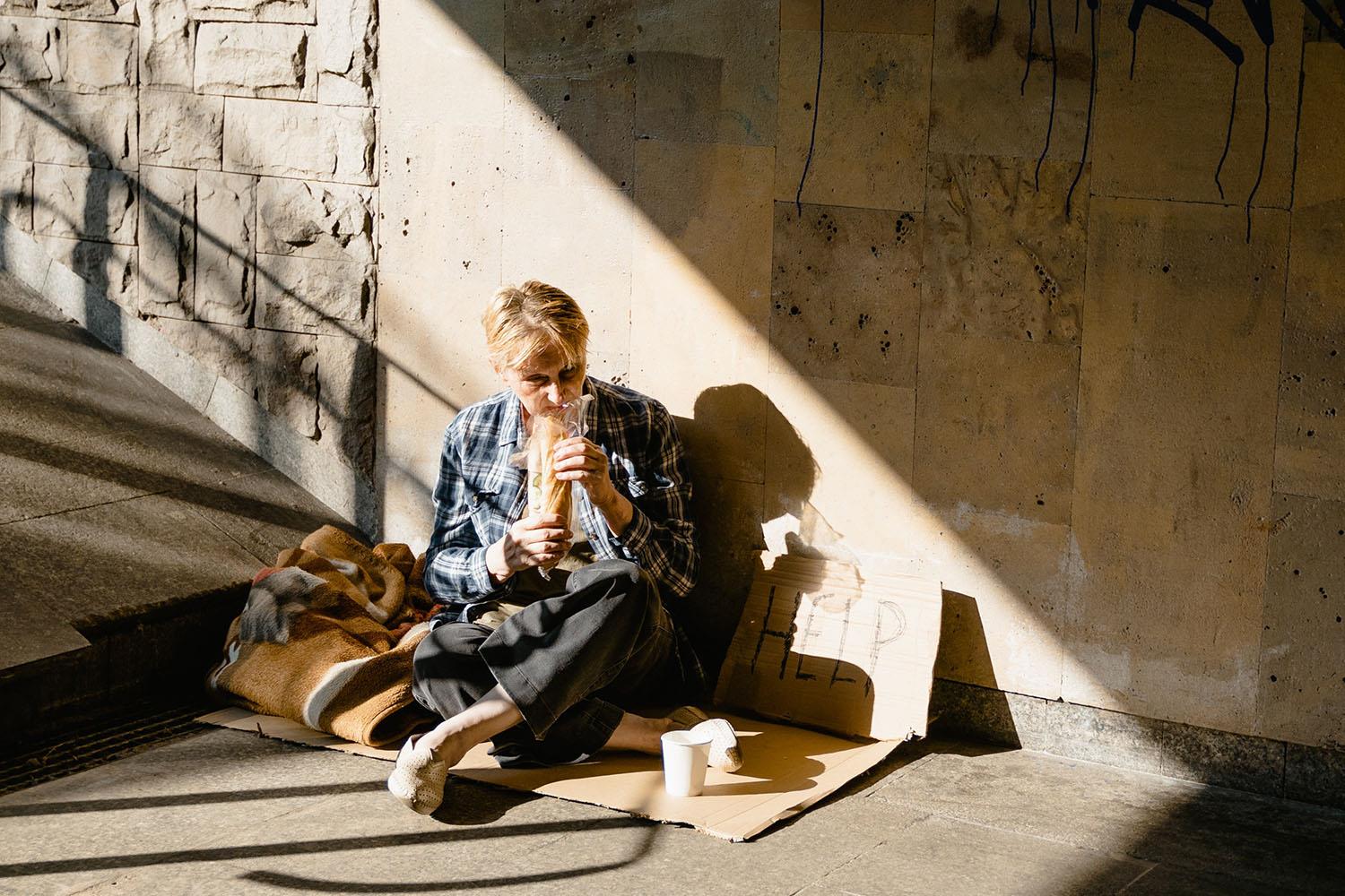 Eine Frau mit kurzen blonden Haaren sitzt auf der Straße und isst ein Brot.
