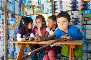 Vier Kinder sitzen an einem Tisch, im Hintergrund eine künstlerische Wand aus Milchtüten.