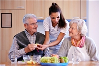 Zwei ältere Menschen bekommen Essen gereicht von einer jungen Frau.