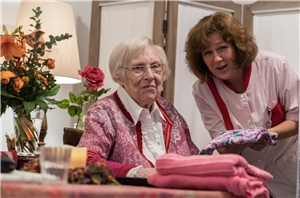 Eine Pflegerin sitzt am Bett einer alten Dame und hält ihre Hand, beide lächeln den Betrachter an.