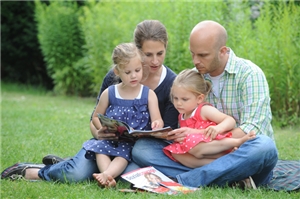 Eine junge Familie mit 2 Kindern lesen ein Buch, auf der Wiese sitzend.