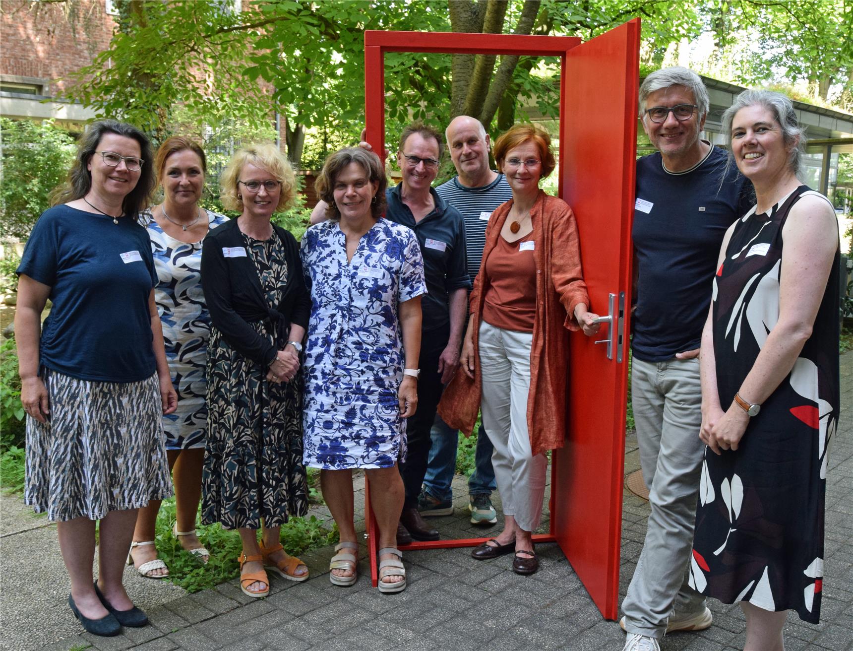 Eine Gruppe von Frauen und Männern steht in, neben und hinter einer frei in einer Parklandschaft stehenden roten, geöffneten Tür.