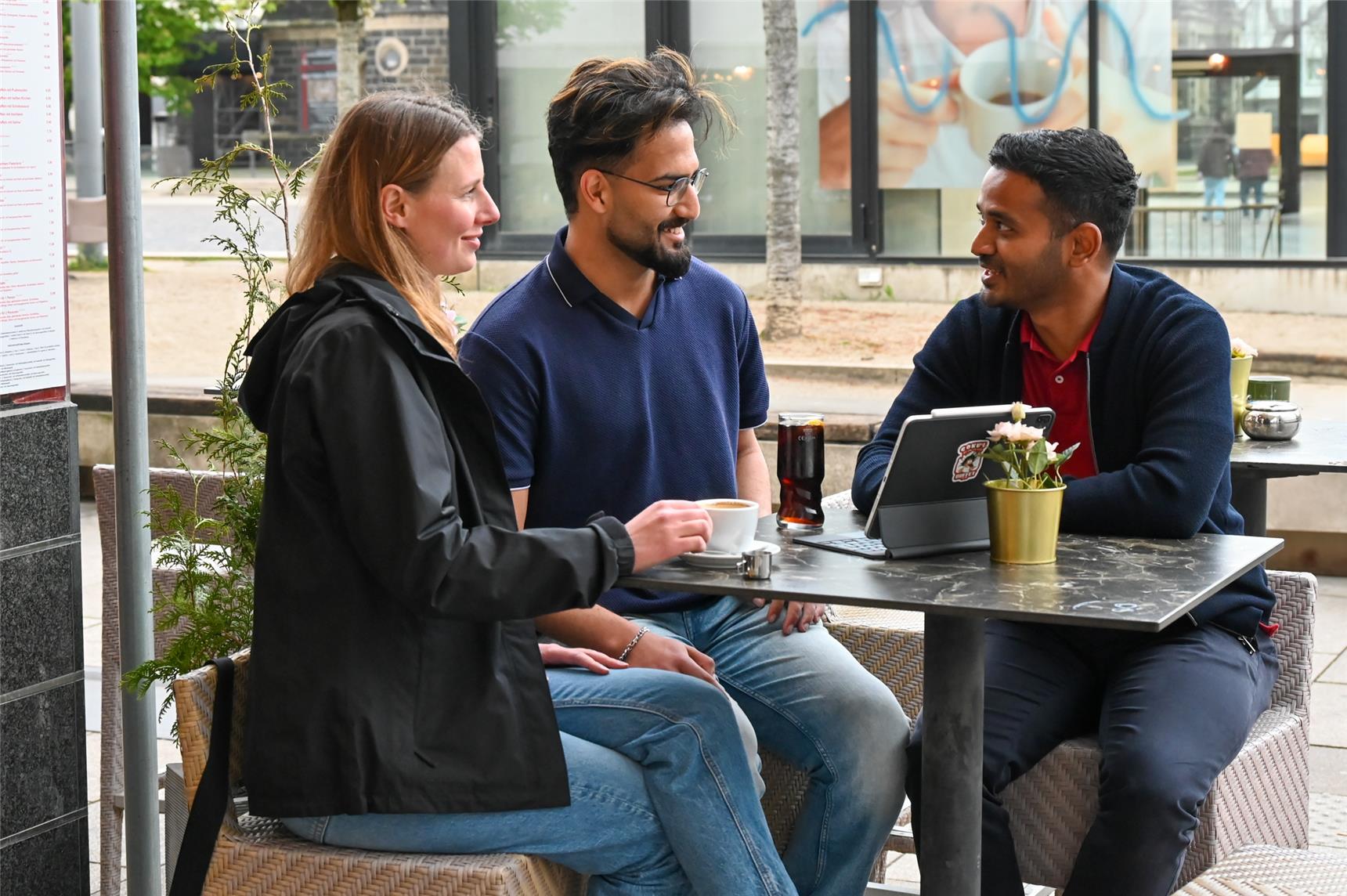 Zwei Männer und eine Frau unterhalten sich an einem Tisch vor einem Café.