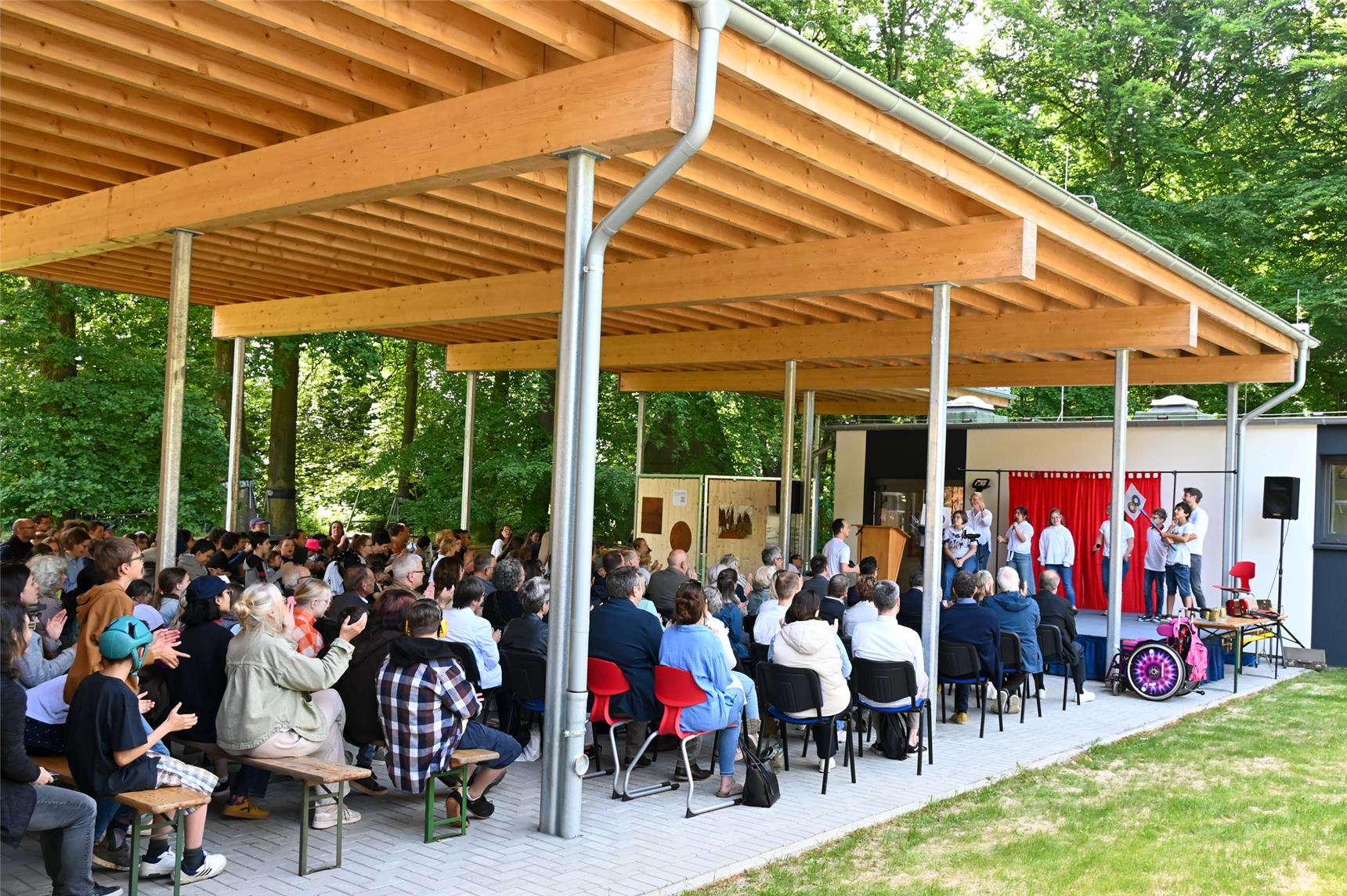 Auf dem Bild sieht man vielen Menschen in Reihe hintereinander sitzend auf Stühlen und Bänken vor einer Bühne. Sie applaudieren. 
