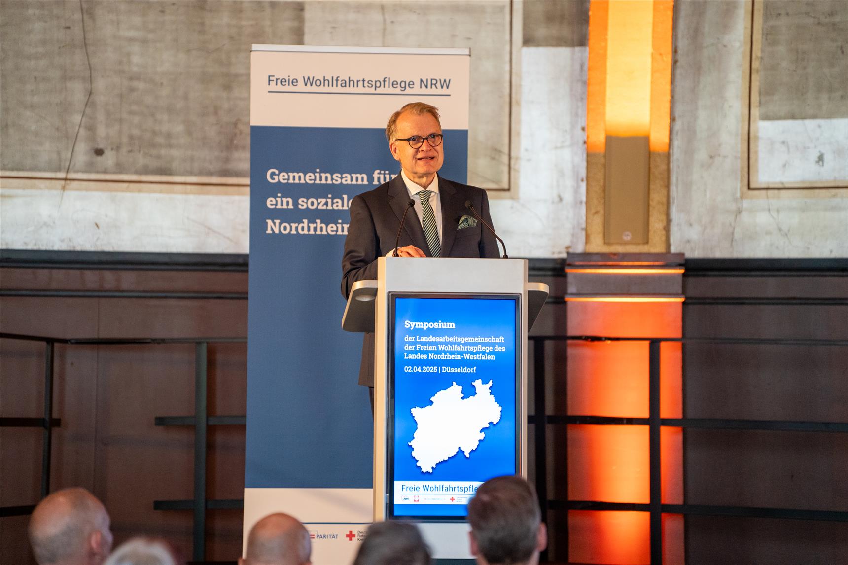 Ein Mann mit Brille an einem Rednerpult, Im Hintergrund ein RollUp der Freien Wohlfahrtspflege NRW, im Vordergrund sind die Hinterköpfe einiger Zuhörer zu sehen.