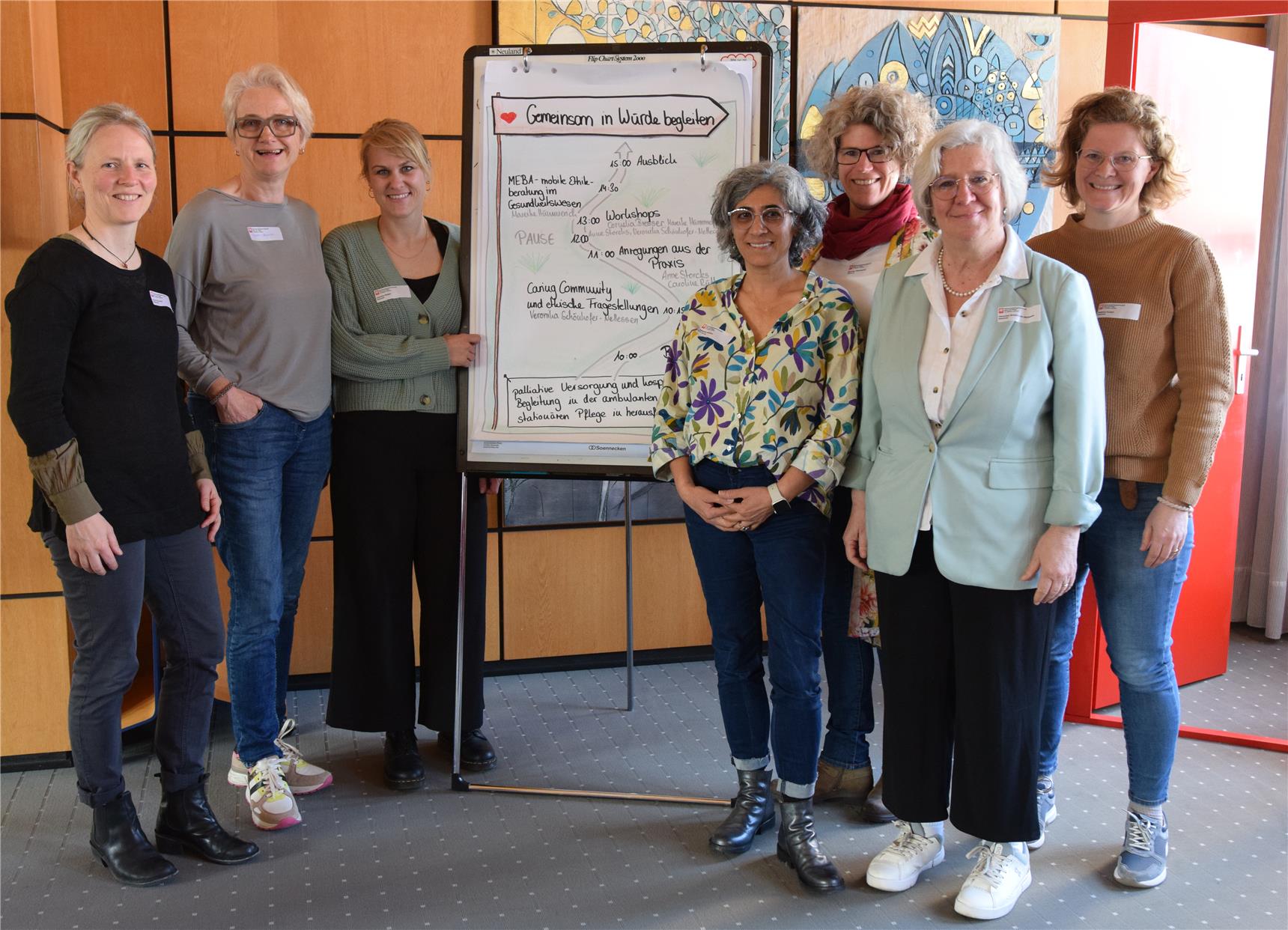 Eine Gruppe von sieben Frauen stehen links und rechts von einem Flip-Chart. Auf dem Titelblatt des Flip-Chart steht: "Gemeinsam in Würde begleiten".