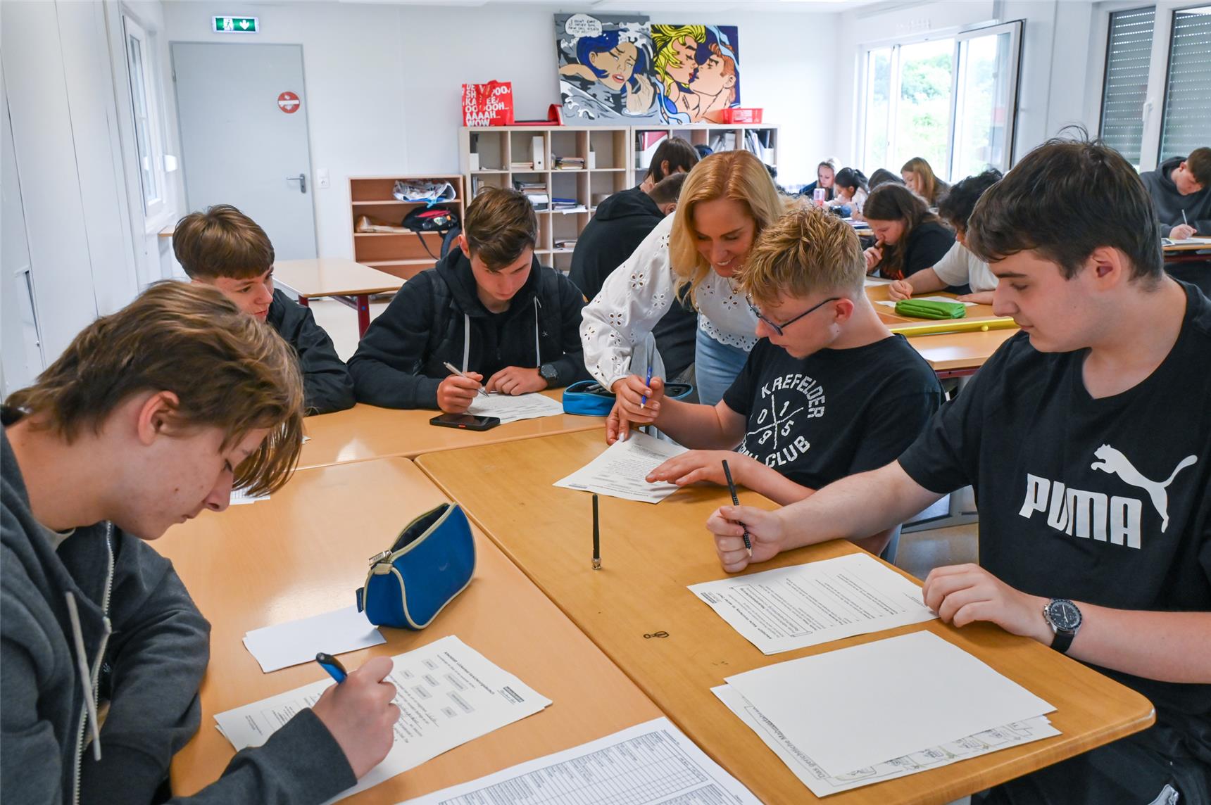 Gezeigt wird ein Klassenraum mit Schülern die in Gruppen um Tische herum sitzen und lernen. Eine Lehrerin gibt dabei Untersützung. 
