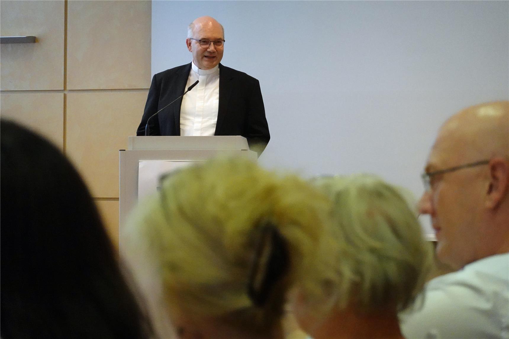 Der Bischof von Aachen, Dr. Helmut Dieser, steht mit Priesterkragen und schwarzem Anzug an einem Rednerpult vor einem weißen Hintergrund. Die Zuhörer sind teilweise im Profil und von den Hinterköpfen her zu sehen.