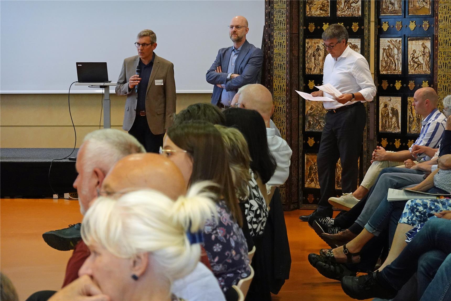 Drei Männer stehen vor einer Zuhörerschaft, die sitzt und zum Teil unscharf im Profil zu erkennen ist. Im Hintergrund steht links vor einer Leinwand ein Stehpult mit einem Notebook darauf, daneben ein religiös anmutendes Kunstwerk, das aus goldfarbenen Darstellungen besteht, die schwarz eingerahmt sind.