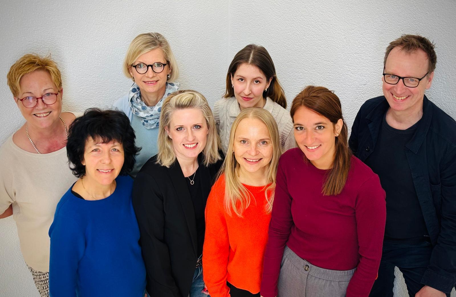 Eine Gruppe von sieben Frauen und einem Mann steht in Zweierreihen vor einem hellen Hintergrund. Das Foto ist von einem leicht erhöhten Standpunkt aus aufgenommen.