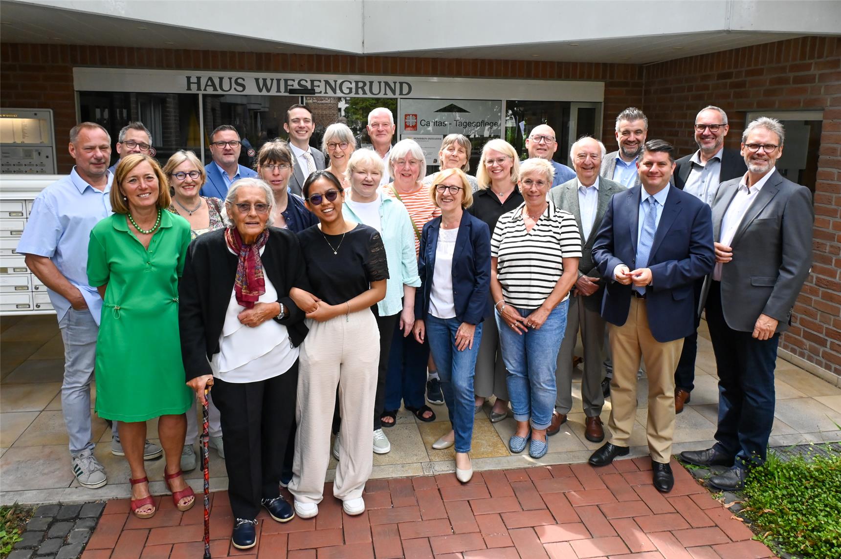 Eine Gruppe von Männern und Frauen steht in Dreierreihe vor einer Caritas-Tagespflege. Vorne links ist eine alte Dame zu sehen, sie ist eine Besucherin der Tagespflege